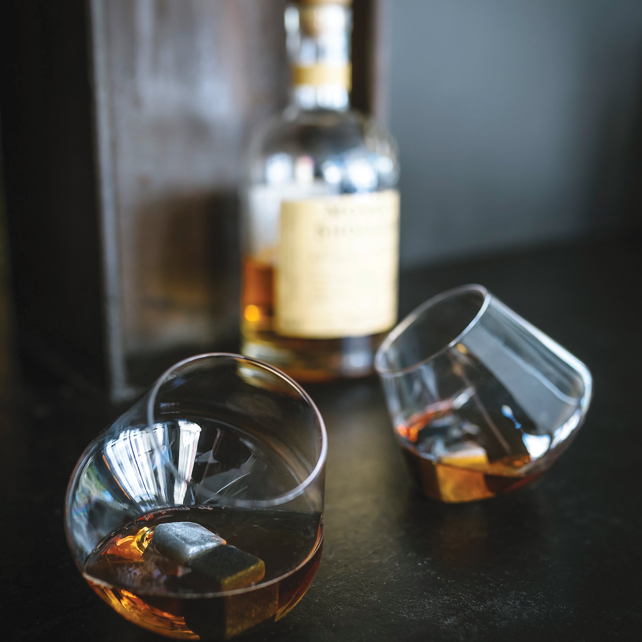 Two Rocking Whisky Glasses with whisky and chilling stones on a dark surface, a whisky bottle behind