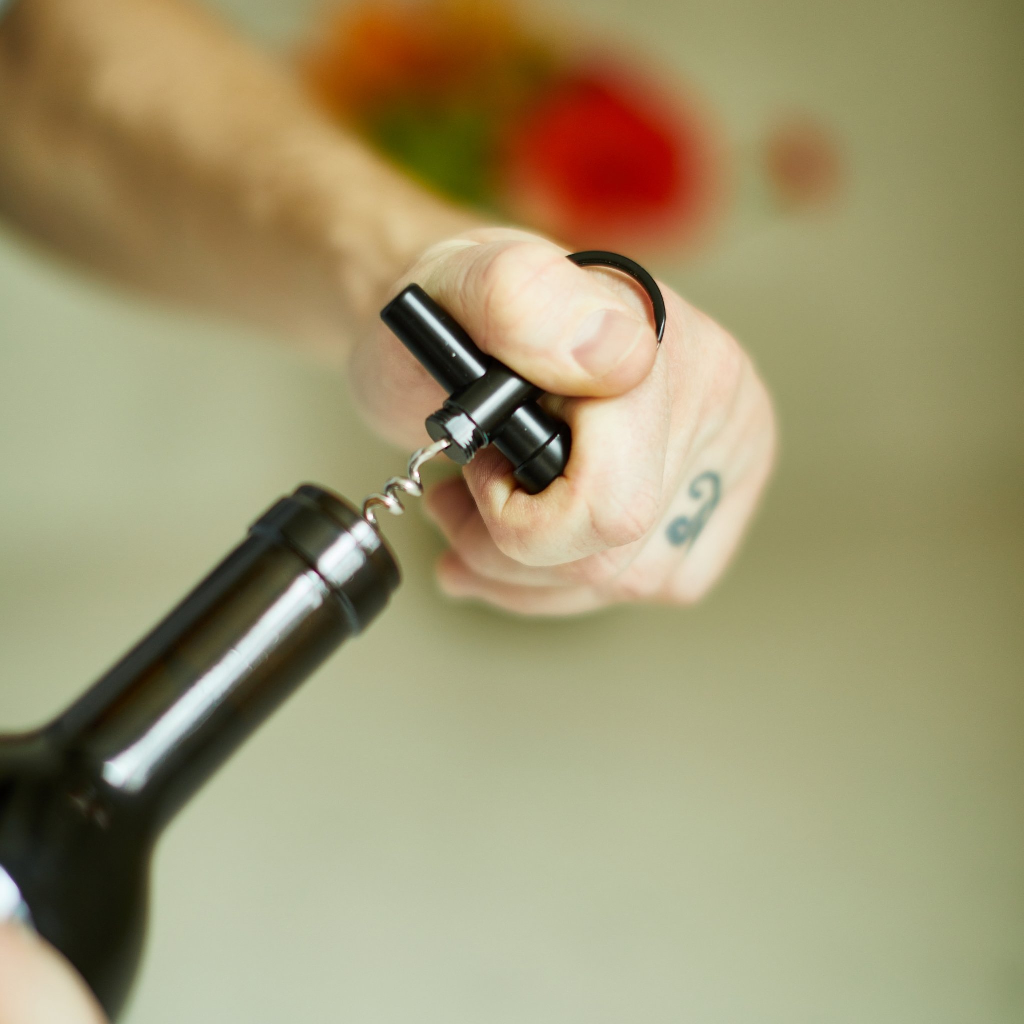 Keychain corkscrew in a fist with its tip at a wine bottle's cork, blurred background behind