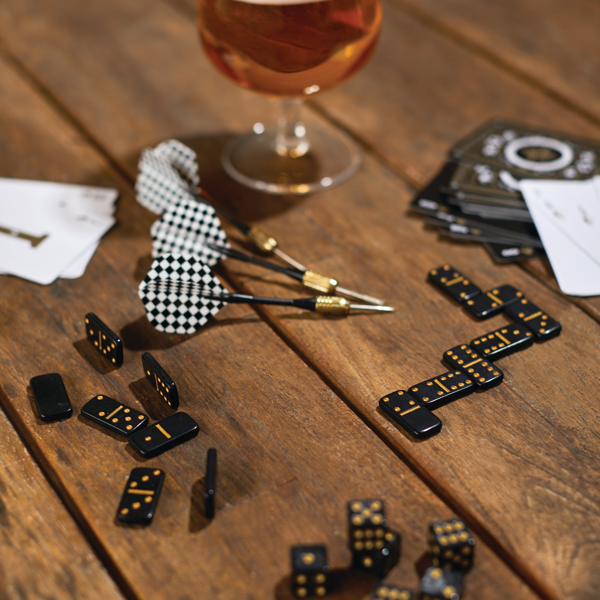 Auf einem Holztisch liegen Darts, Dominosteine, Würfel und Karten neben einem Glas Bier, mitten im Spiel
