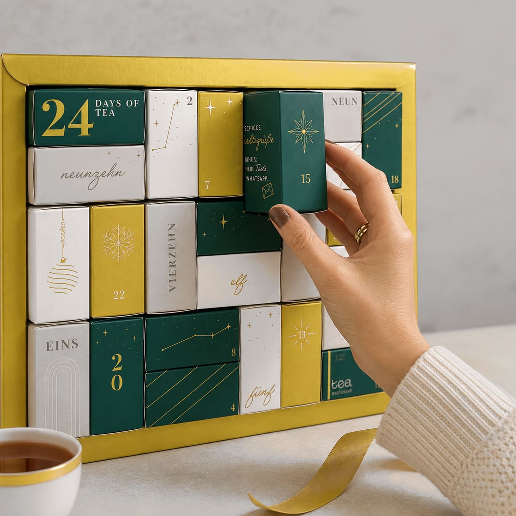 Hand zieht eine grüne Schachtel aus dem Tee-Adventskalender, davor eine Tasse Tee