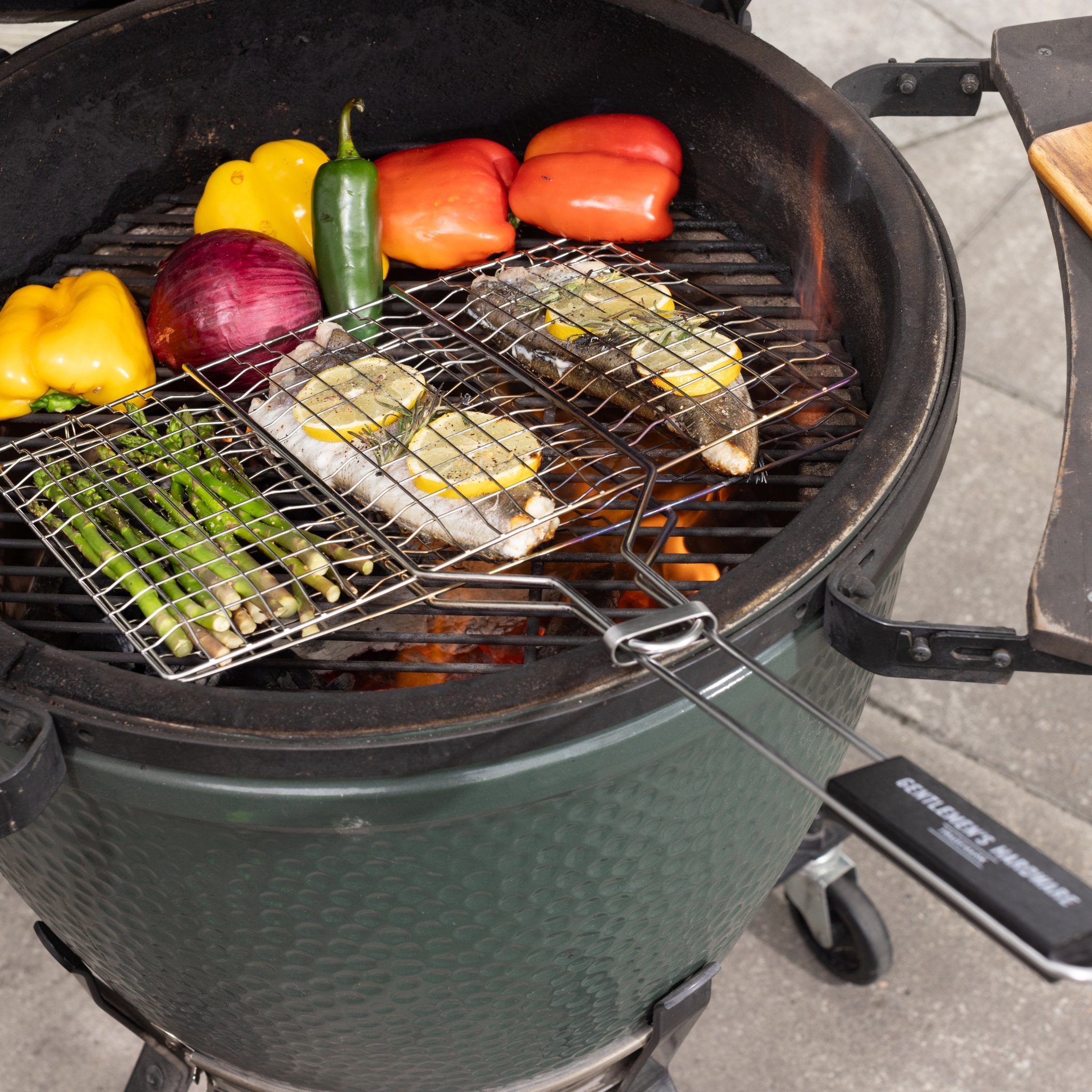 Cestini per barbecue visti dall'alto su una griglia in ceramica verde, lungo manico con logo in primo piano, verdure sulla griglia