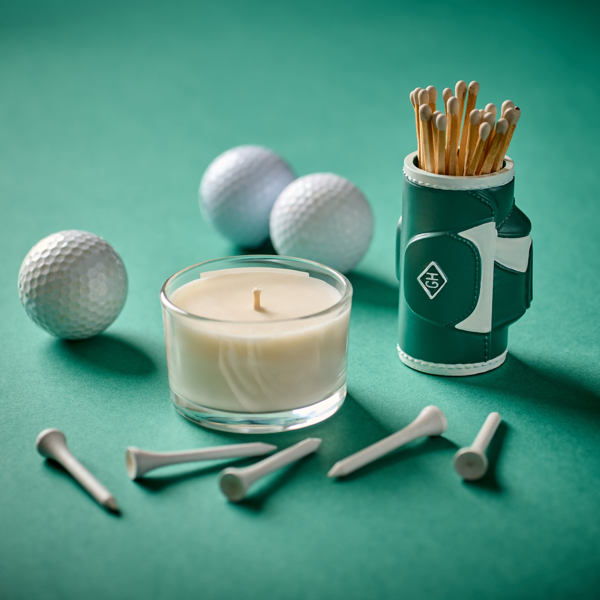 Golf bag match holder beside a candle, golf balls and tees on green