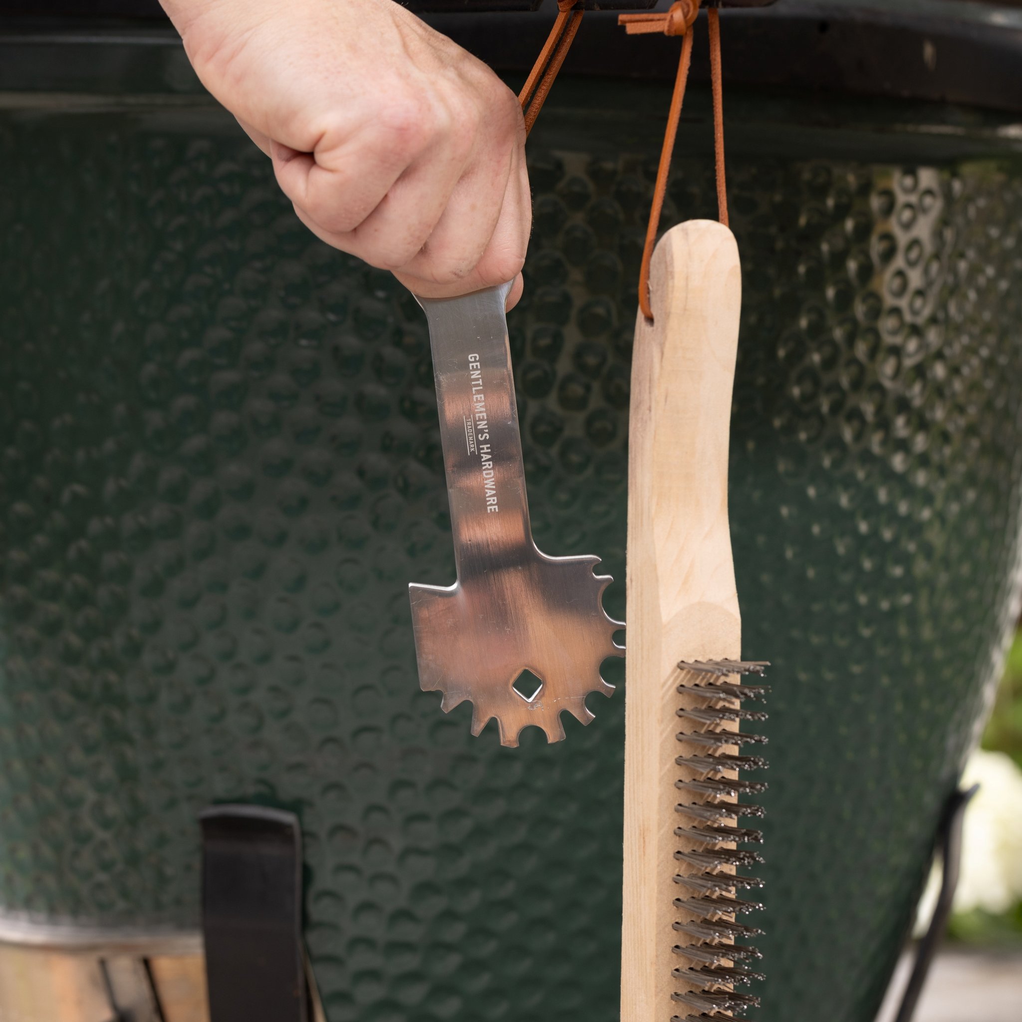 Une main tient le grattoir du kit pour l'entretien du barbecue, la brosse métallique pend à côté devant un gril en céramique vert