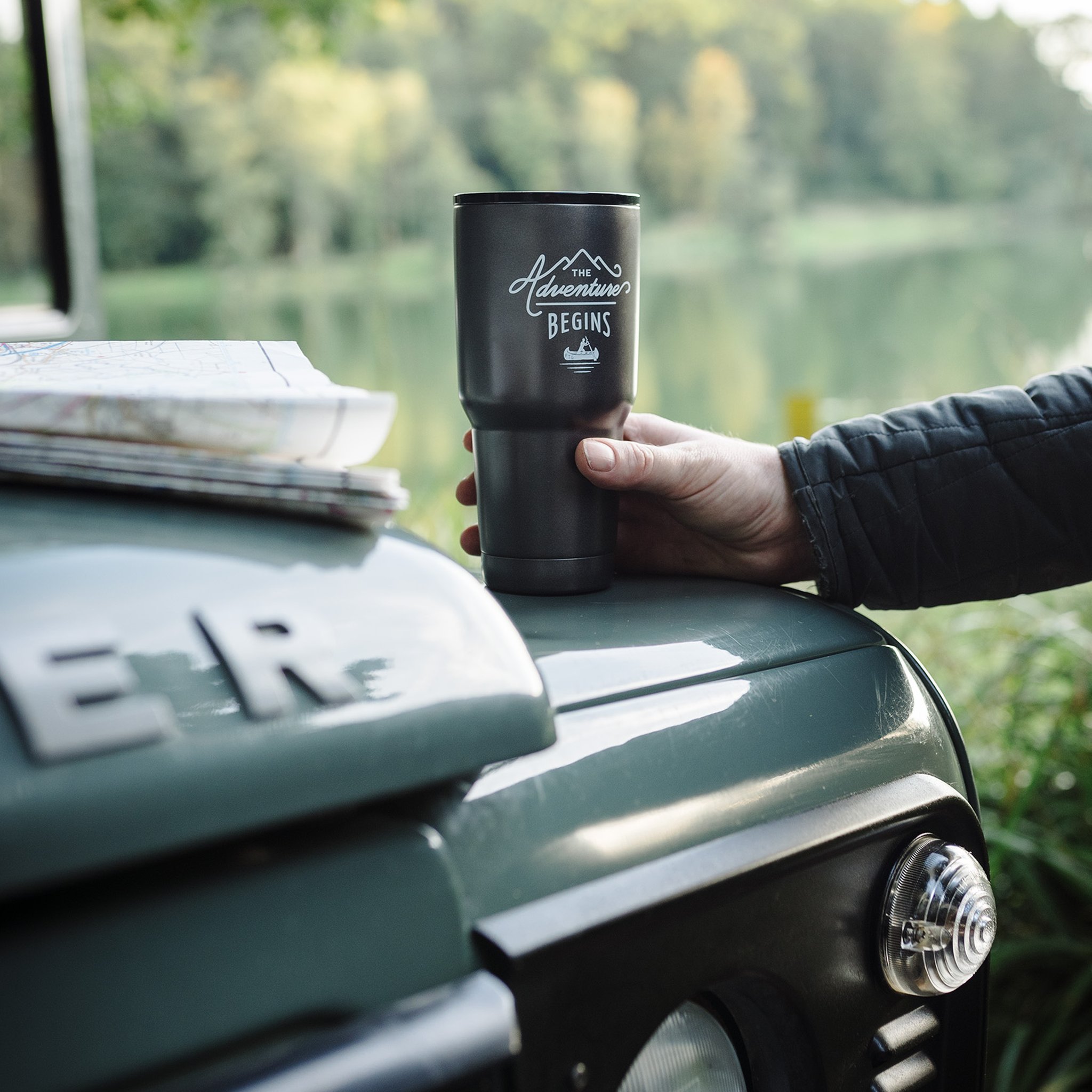 Hand hält die Reise-Kaffeepresse auf der Motorhaube eines Gelaendewagens, daneben Landkarten und ein See