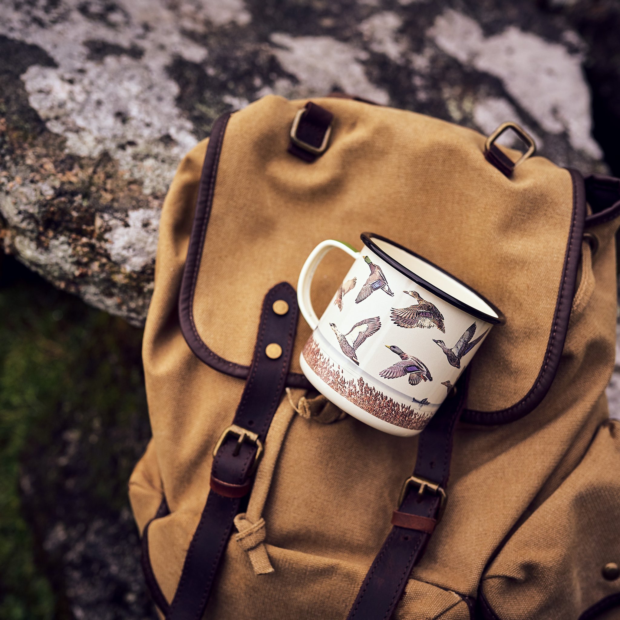 Enten Emaille-Tasse liegt draussen auf einem beigen Rucksack mit Lederriemen, im Hintergrund ein moosiger Fels