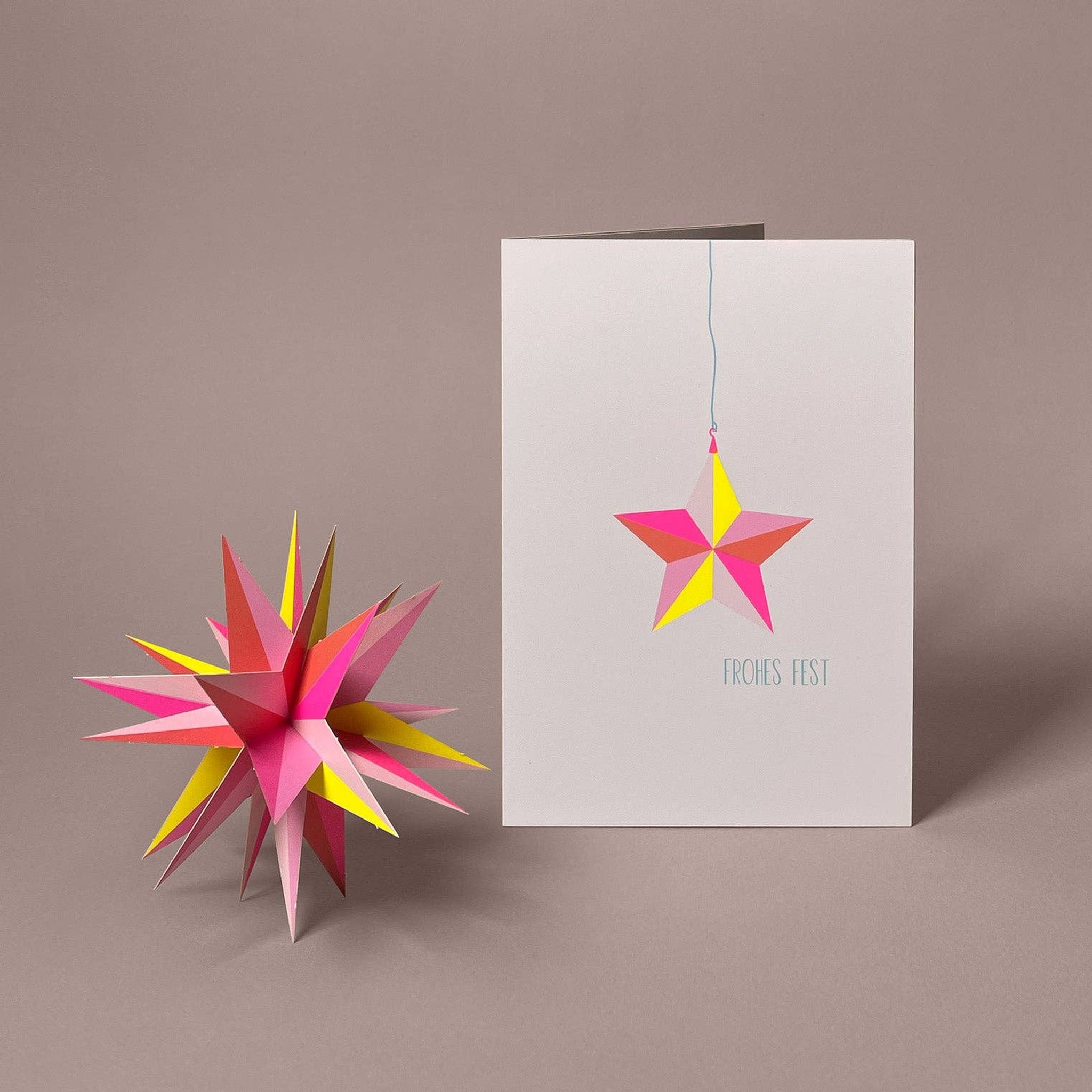 Gefaltetes 3D-Modell Winterstern in Neonpink und Neongelb neben der Weihnachtskarte «Frohes Fest» von Faltmeister