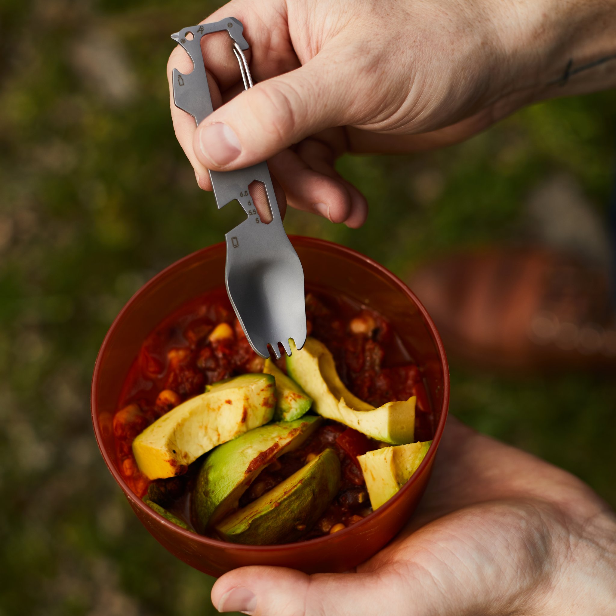 Gros plan d'une main soulevant la fourchette multifonctions d'un bol de chili et d'avocat, verdure au fond