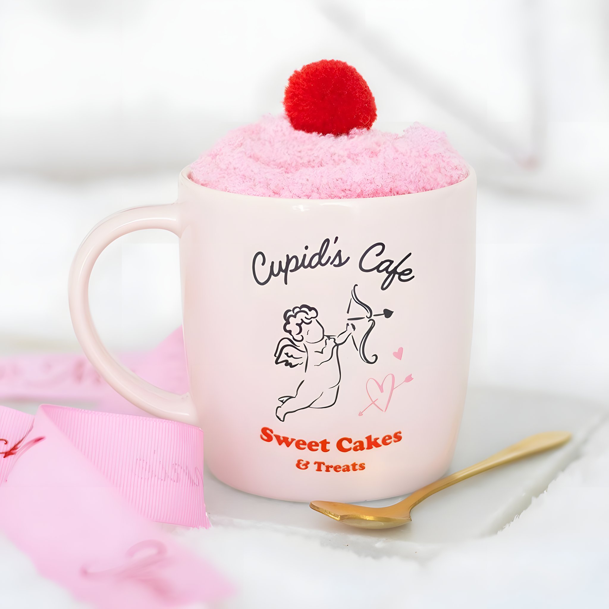 Tasse rose Cupid's Café avec chaussettes roulées et pompon sur un lit