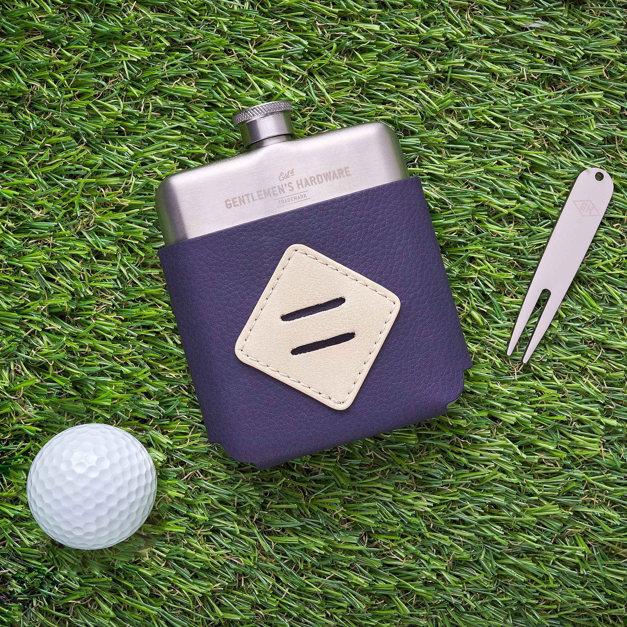 Hip flask in a blue pouch lying on artificial turf, with the divot tool and a white golf ball beside it