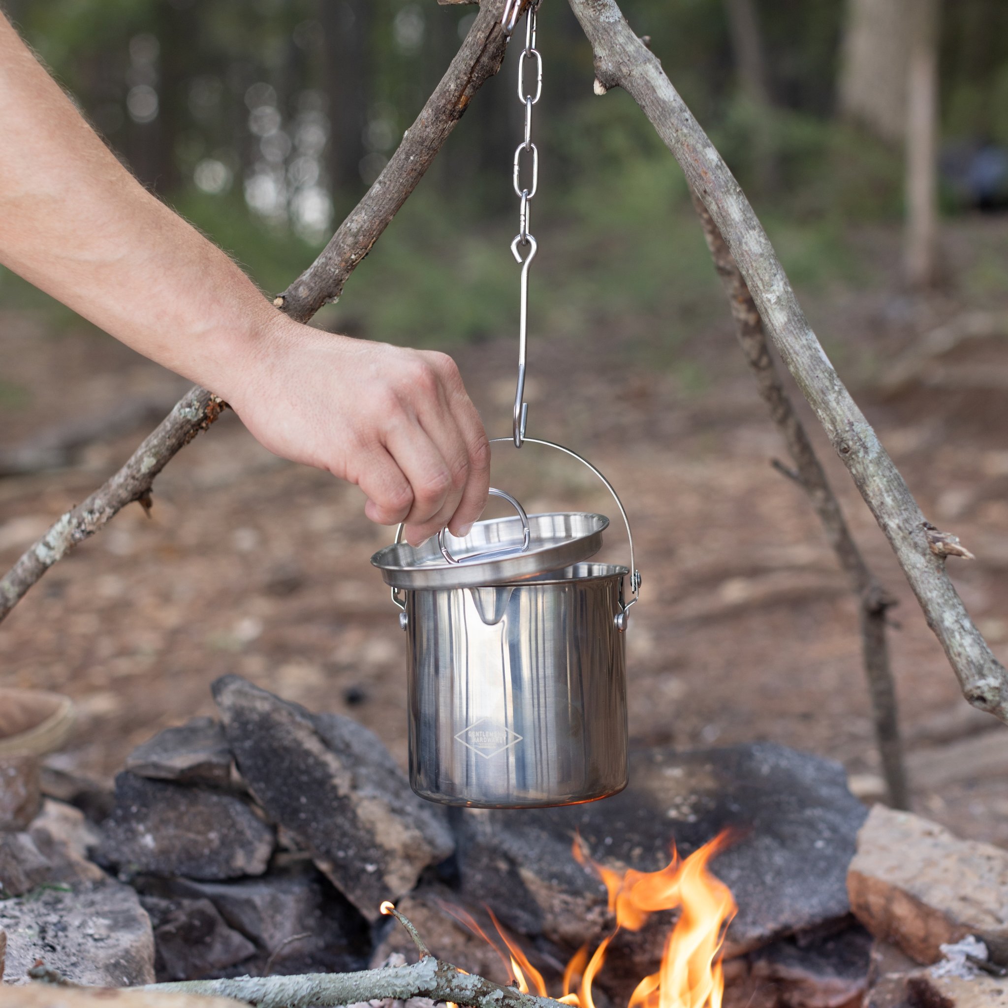 A hand lifts the lid of the Campfire Cooking Pot as it hangs from a tripod over a fire in the woods