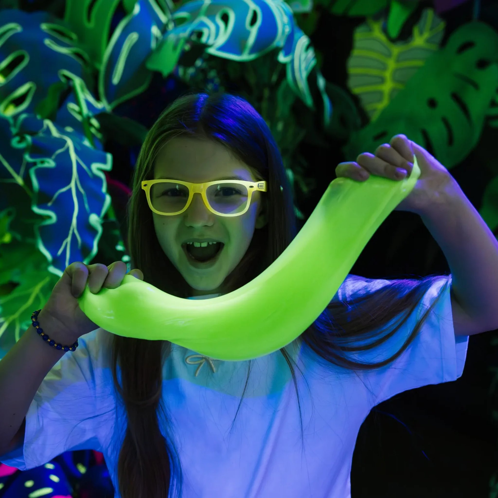 Ragazza con occhiali gialli che allunga lo slime verde luminoso sotto la luce nera