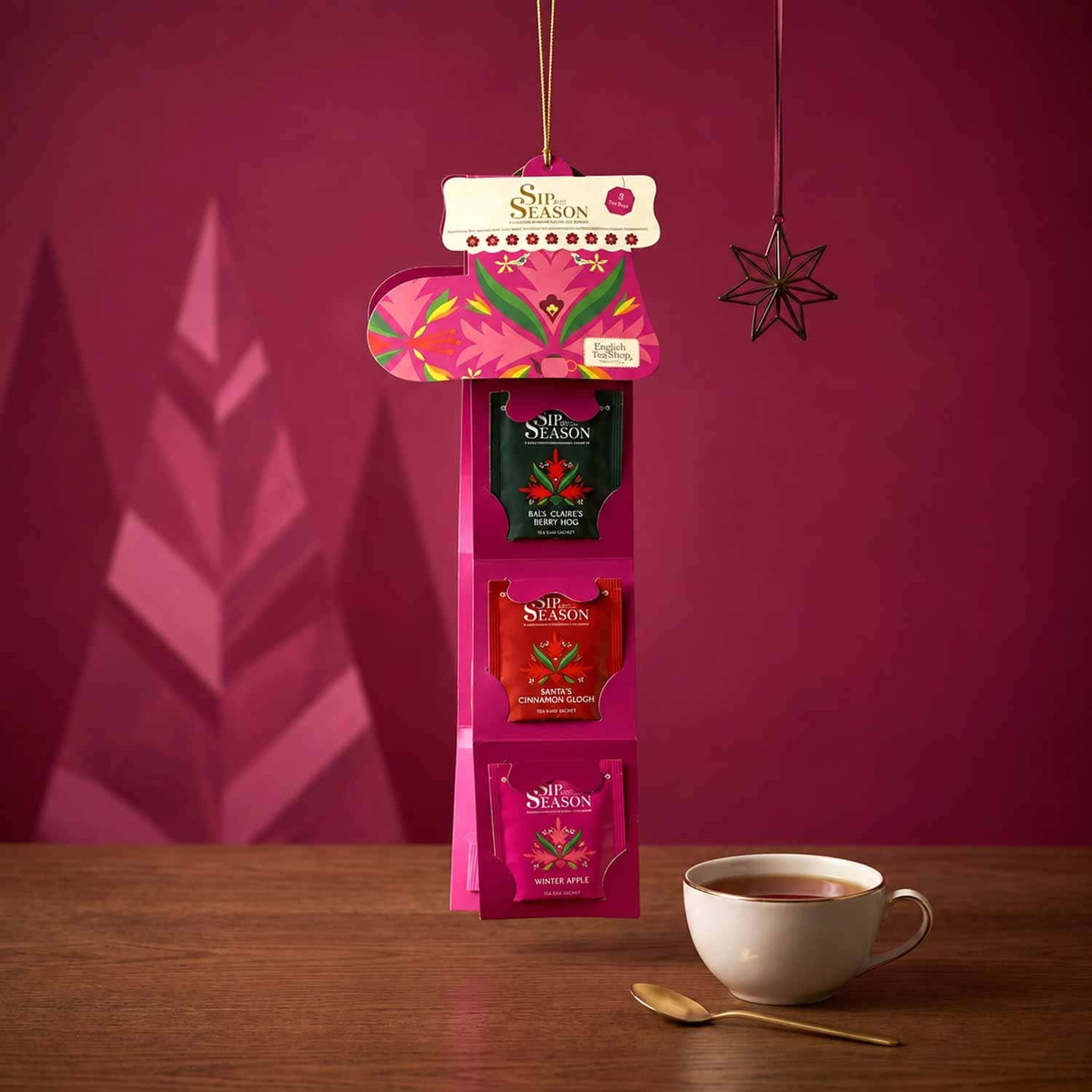 Botte de Noël dépliée suspendue devant un mur framboise, montrant trois sachets, tasse de thé devant