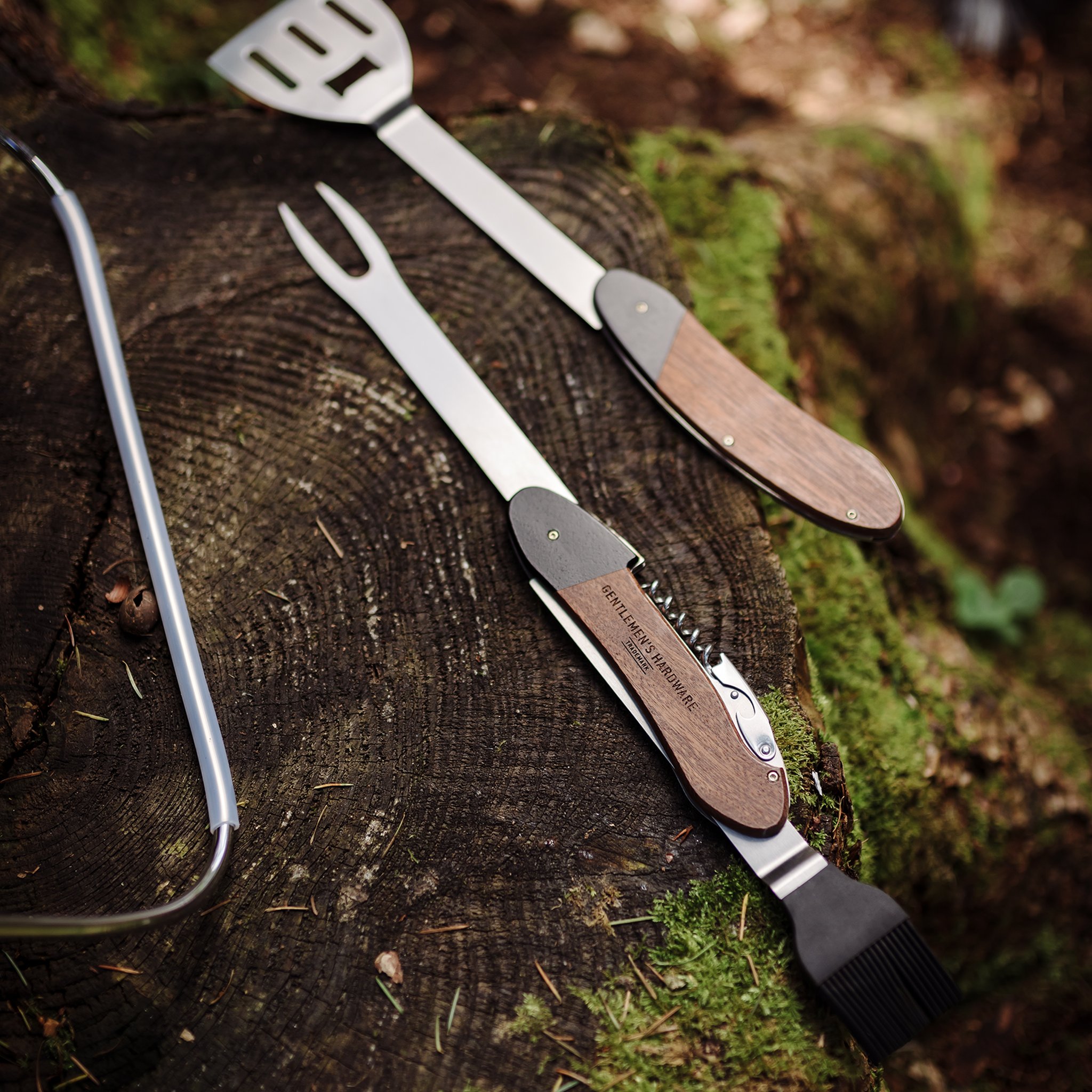 Unfolded BBQ Multi Tool resting on a mossy tree stump in the woods, with a metal handle at the left edge