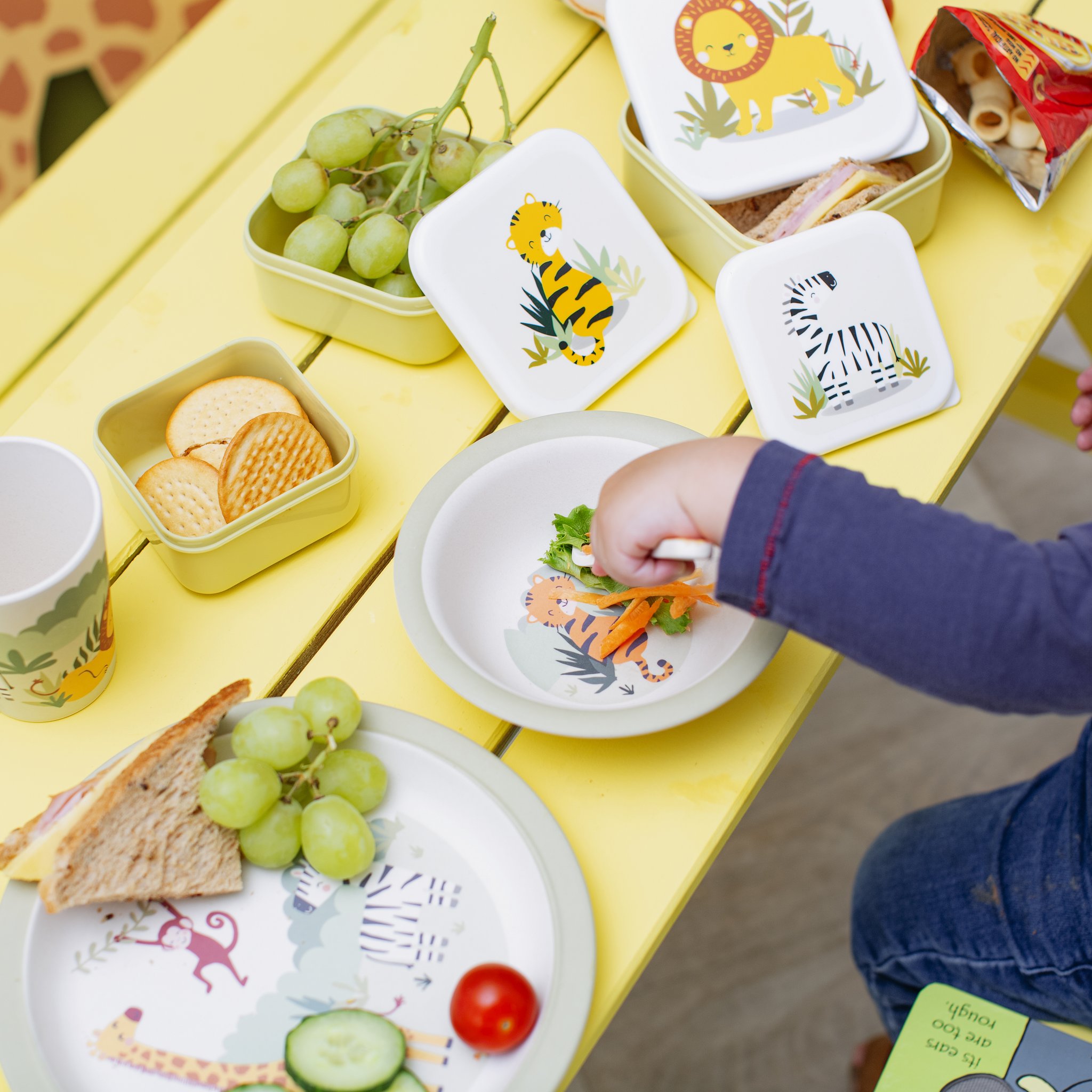 Stoviglie per bambini Savannah Safari con uva e portapranzo su tavolo giallo, vista dall'alto