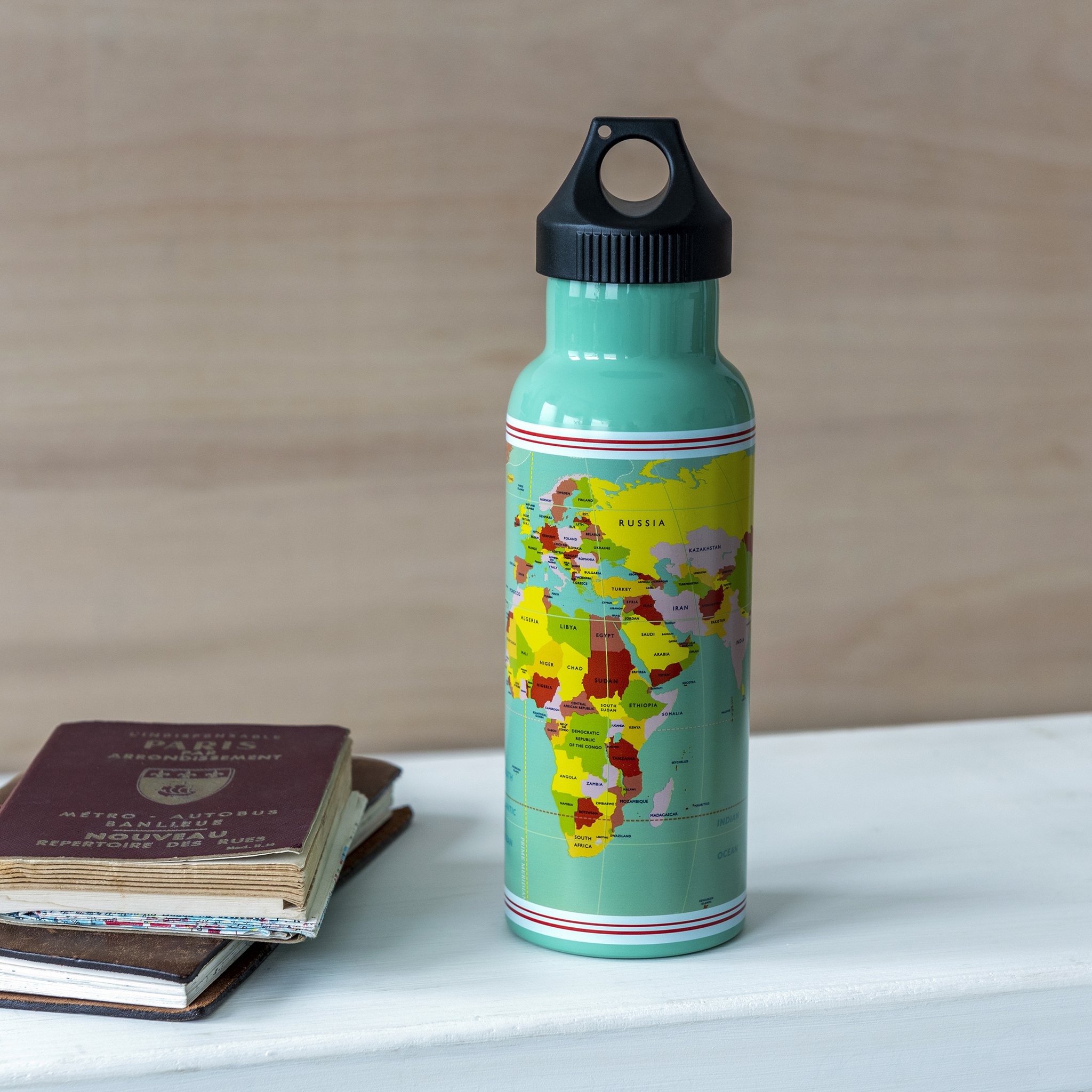 Weltkarten-Trinkflasche aus Edelstahl neben Reisepass und Notizbuch auf der Bank