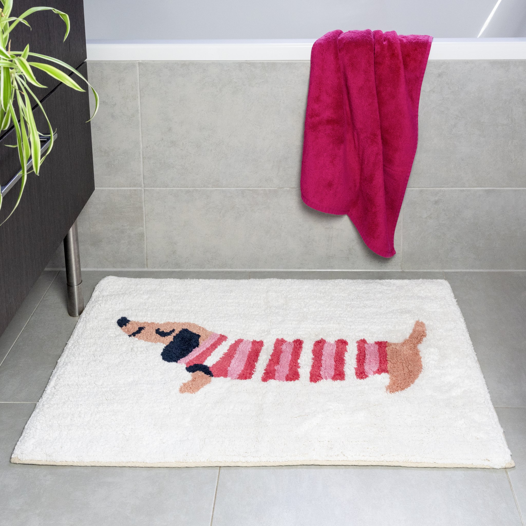 Tappetino da bagno bassotto davanti alla doccia con asciugamano rosa e pianta verde