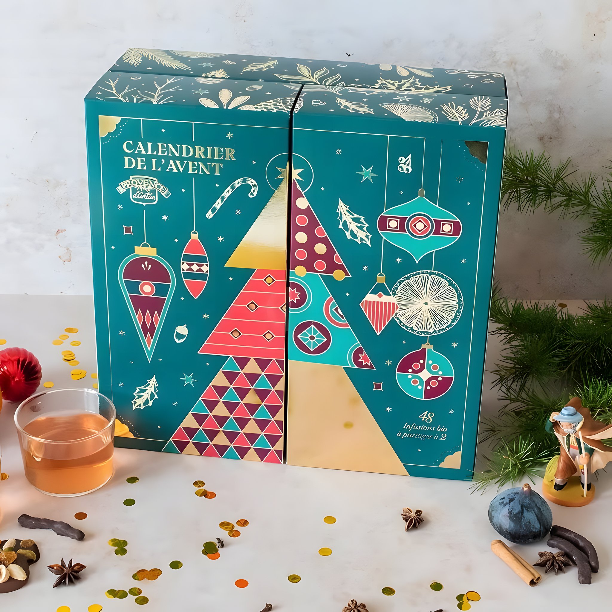 Geschlossene petrolgrüne Kalenderschachtel mit Weihnachtsbaum-Motiv, daneben eine Tasse Tee