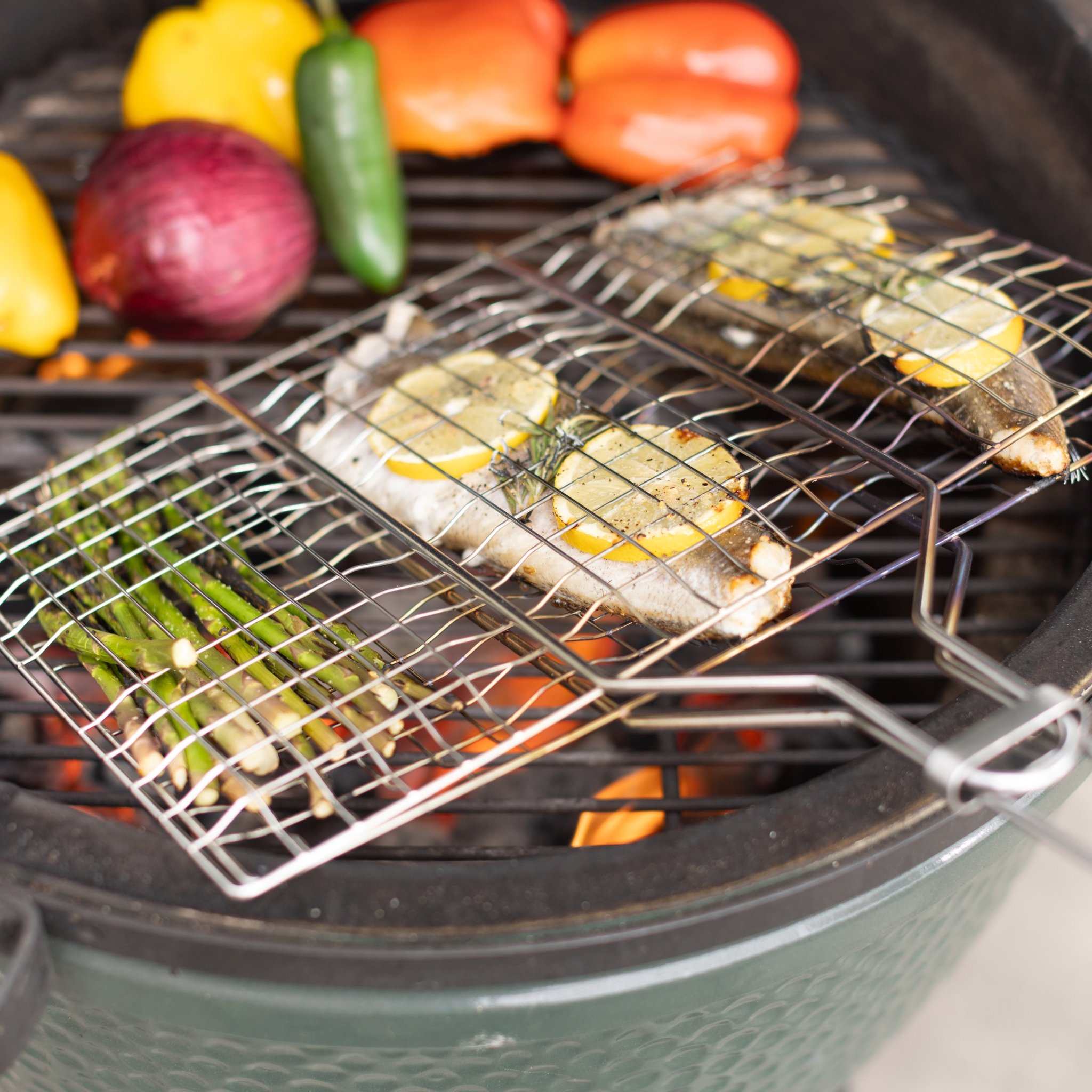 Primo piano dei cestini per barbecue sulla griglia sopra la brace, con pesce, limone e asparagi, cipolla rossa sullo sfondo