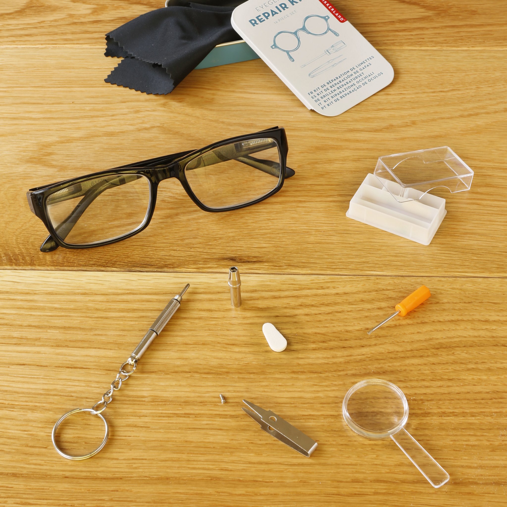 Kit riparazione occhiali disposto su legno: occhiali, cacciavite, lente, ricambi e scatola