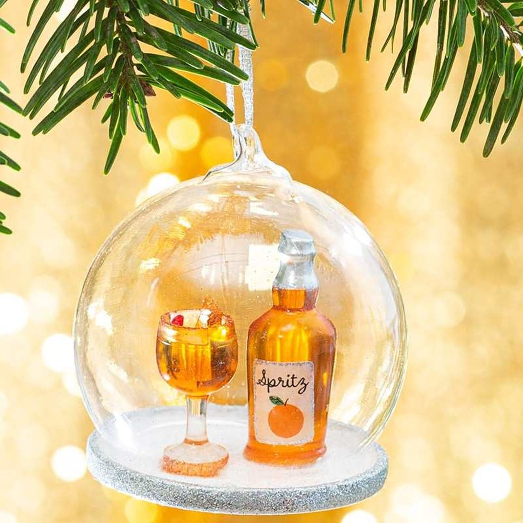 Boule de Noël cocktail Spritz Sass & Belle sur une branche de sapin, dôme en verre avec bouteille et verre